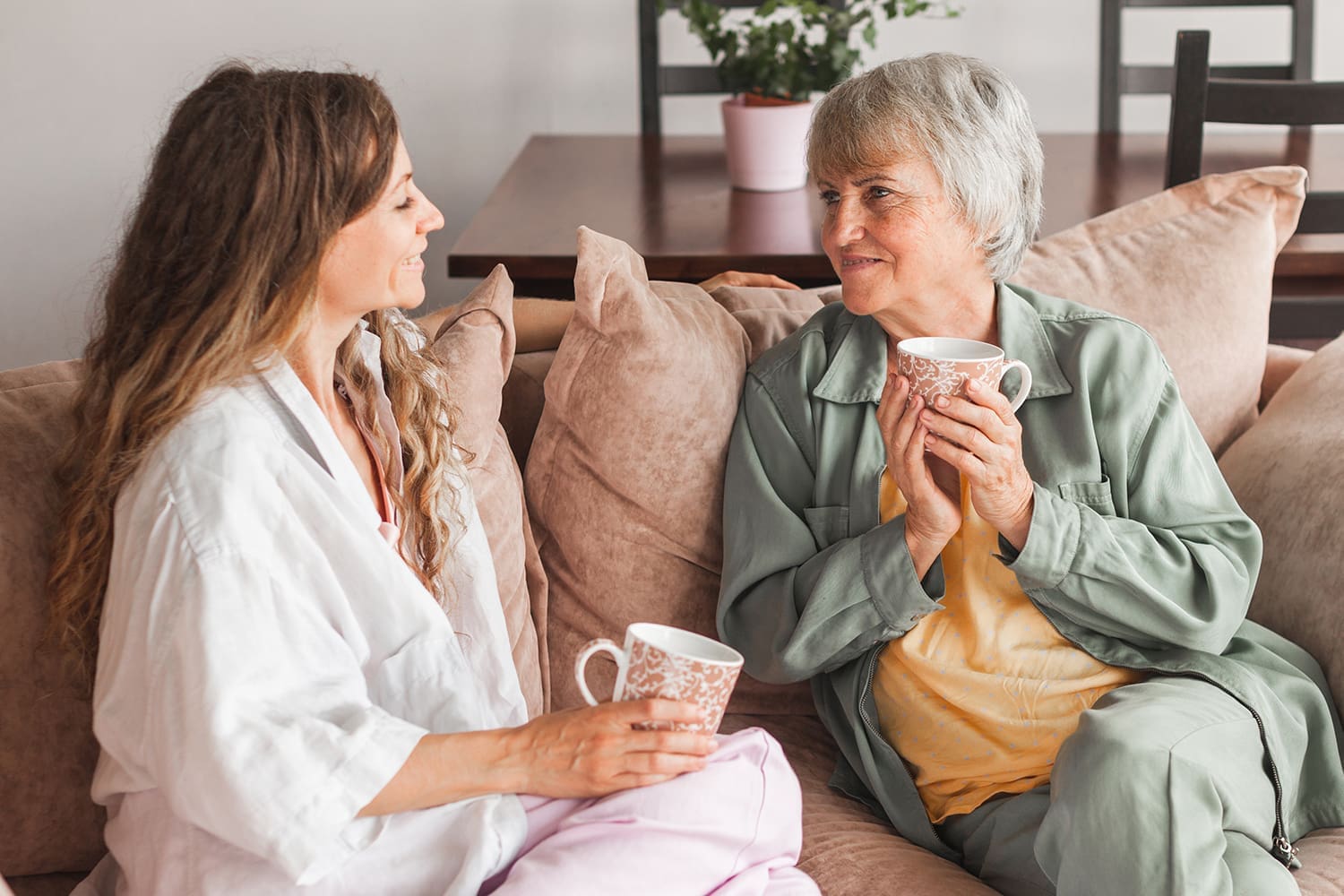 Two women holding mugs, sitting on a couch talking.